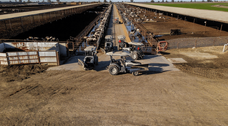 A sequence of tractor images of the gray and teal (tractor battery) MK-V dairy tractor shows how tractor makes a U-turn at the end of the dairy aisle, without a driver for autonomous dairy feed pushing. Foreground is brown dirt road. 