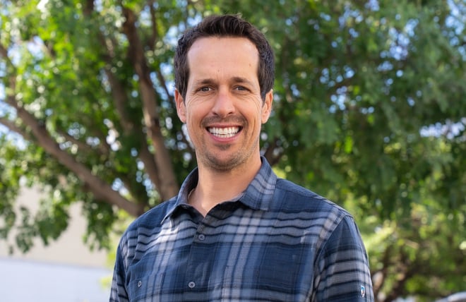 Smiling image of a man pictured from the chest up wearing a blue plaid shirt. He has brown hair, olive-pale skin and slight beard growth. It