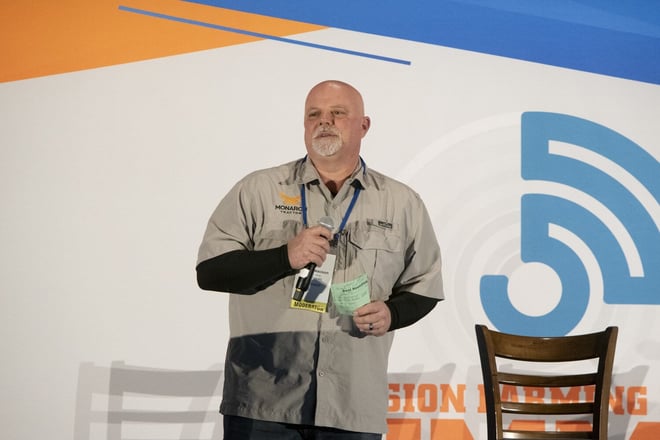 Man, bald, gray goatee wearing in khaki button up shirt with Monarch Tractor brand is speaking on a stage. 