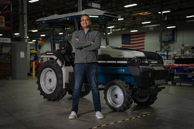 Monarch Tractor CEO, Praveen Penmetsa, stands in front of MK-V electric tractor with U.S. flag in background. Smiling, black hair, arms folded, wearing jeans, white sneakers, and a gray jacket. 